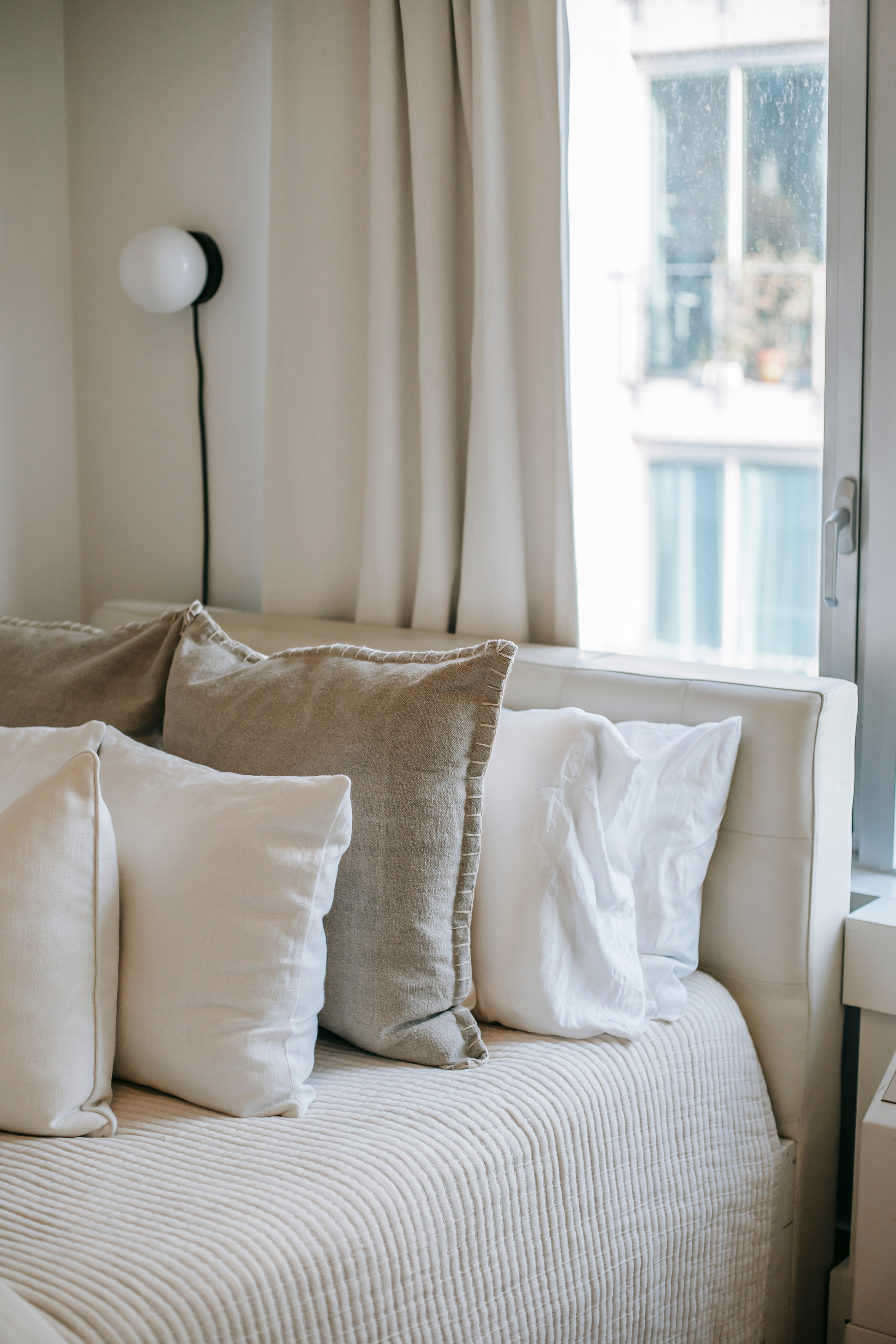 A cozy bed with white sheets, linen coverlet, and airy pillows in front of a bright window.